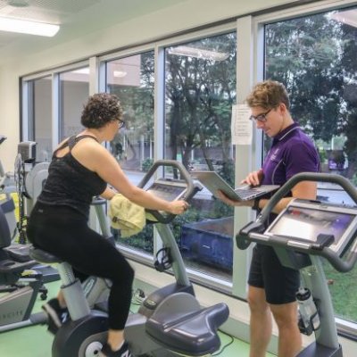 Woman on exercise bike while man stands nearby with laptop. Supplied.